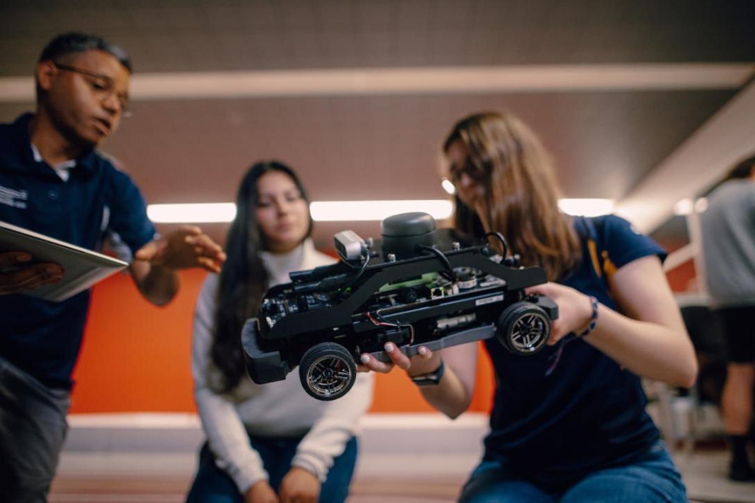 One Kettering student holds a small autonomous vehicle while another watches. A Kettering professor gives instructions.